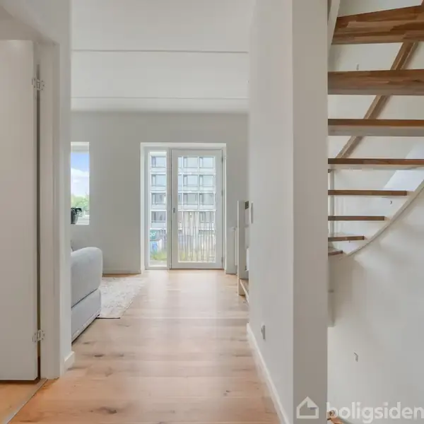 Trappe fører opad ved siden af en lys gang med træparket og indgang til en stue med åbne døre til en balkon. Lysindfald fra vinduer.