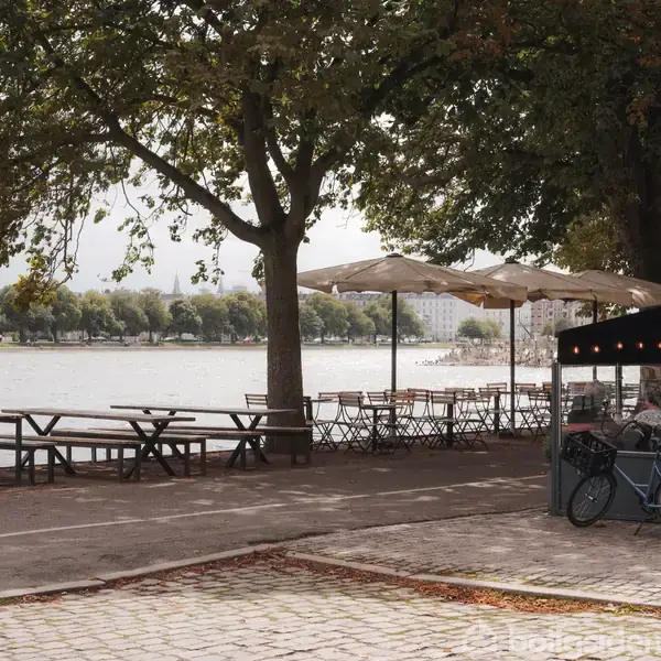 Cykel parkeret ved caféområde med borde og parasoller, der vender mod søen. Træer omgiver området, og der er gåsti langs vandet. Skilt viser "La Vague".