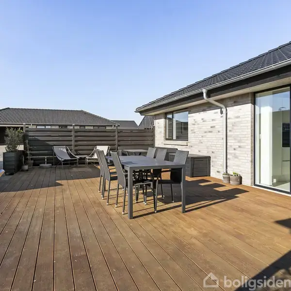 En udendørs træterrasse med et spisebord og stole står foran et moderne hus med store vinduer. Terrassen er solrig og omgivet af hegn, hvilket skaber en privat atmosfære.