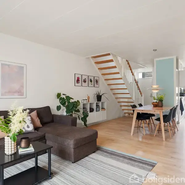 En stue med en mørk sofa står ved siden af et sort sofabord med blomster. En åben trappe fører til næste etage. Spisebord med stole står i nærheden. Tre billeder hænger på væggen.
