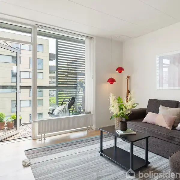 Stue med grå sofa, et lille sofabord og gulv-til-loft-vinduer åbner til en lille altan. Lyse vægge dekoreret med kunst og rød vægbelysning.