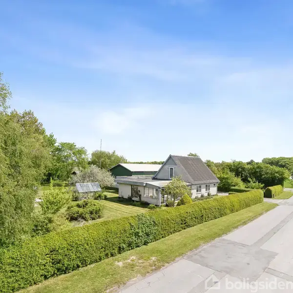 Et hus med skråt tag står omgivet af en frodig have omkranset af buske. En vej løber langs huset, og træer ses i baggrunden under en klar, blå himmel.