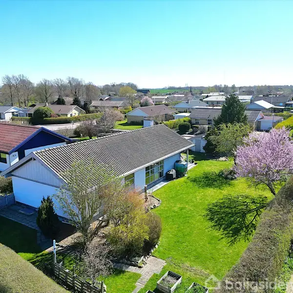 Et én-etagers hus med skråtag står i en grøn have med blomstrende træer. Omgivet af en række andre huse i et forstadskvarter under en klar blå himmel.