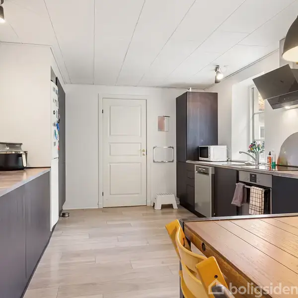 Køkken med mørke træskabe og bordplader, kaffemaskine på bordet. En spiseplads med træbord og gule stole. Lyse vægge, hvid dør, stor loftslampe, planter som dekoration.