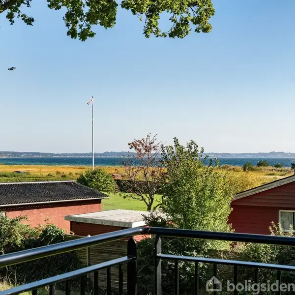 Rækværk står stille på en terrasse med udsigt over huse og træer, mens en flagstang ses i baggrunden nær en sø eller hav.