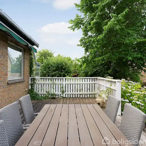 Et rektangulært træbord omgivet af kurvestole står på en terrasse. Ved siden af er et gult murstenshus. Baggrunden viser hegn, træer og en blå himmel.