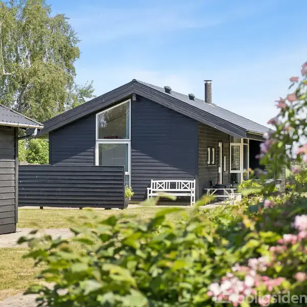 Et sortmalet sommerhus med store vinduer står i en have med grønne buske og blomster. Et solrigt, klart vejr fremhæver den naturlige skønhed i haven omkring huset.