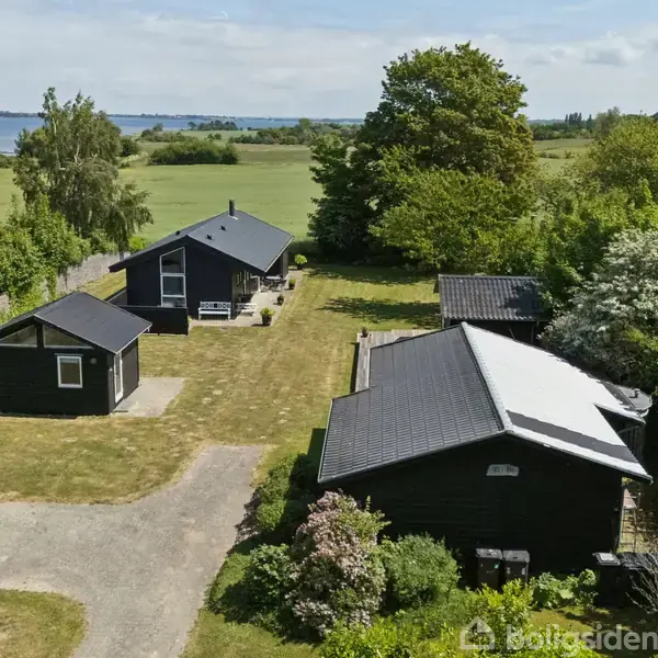 Tre sorte huse står på en græsplæne omgivet af grønne træer og buske. I baggrunden er der udsigt til marker og hav.
