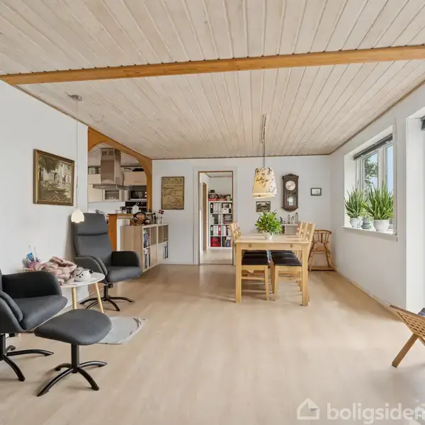 En stue med to sorte lænestole på venstre side står stille. Et spisebord med stole i lyset fra store vinduer. Rummet har lyse trægulve og vægge med kunst.