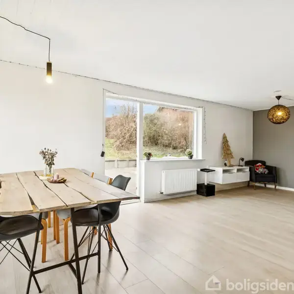 Et lyst rum med et træbord og sorte stole. Sollys strømmer ind gennem et stort vindue, der vender mod grønne omgivelser. En moderne lampe hænger fra loftet.