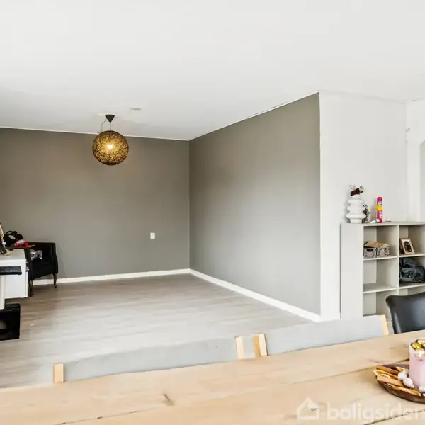 Et rummeligt, moderne stueområde med lysegrå vægge og trægulv. Et rundt, dekorativt lys hænger fra loftet. En spisebordkant med dekoreret bakke ses i forgrunden, mens vinduer med små planter er til venstre.