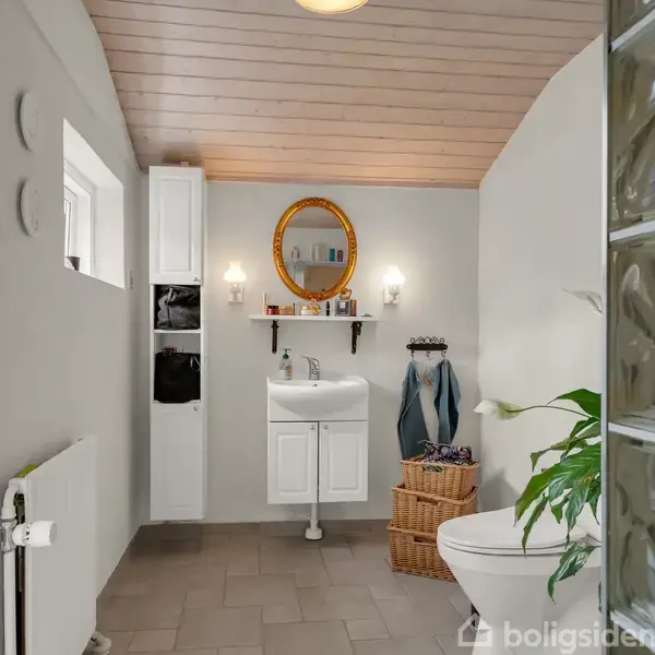 Badeværelse med vask under et rundt spejl, omgivet af væglamper. Kurv og håndklæder til højre. Flisegulv og træloft. Grøn plante ved siden af toilettet.