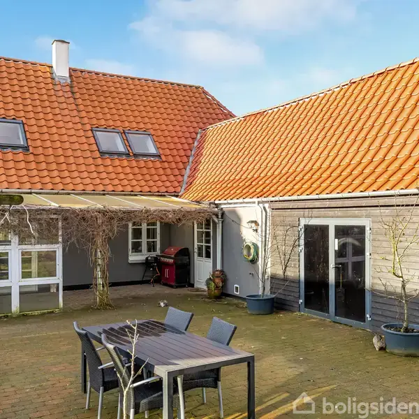 Et bord med fire stole står på en flisebelagt terrasse foran et hus med rødt tegltag. Huset har store vinduer, og omgivelserne er rolige med træer og græsplæner.