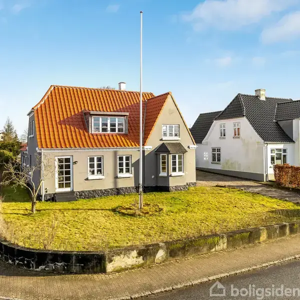 Et grå hus med rødtag står på en hjørnegrund med velplejet græsplæne og en flagstang. Ved siden af findes et hvidt hus. Vejen strækker sig langs husene.