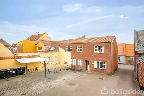 Et rødt murstenshus står stille i en gårdsplads omgivet af andre farvede bygninger. Et udendørs område med borde og stole er synligt ved siden af huset.