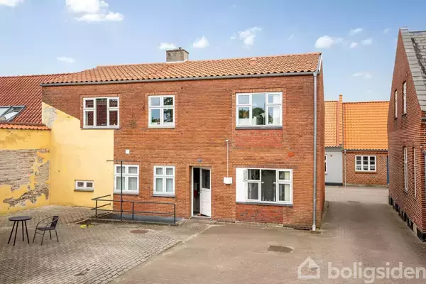 Rødt murstenshus med flere vinduer står i en gårdsplads. En sort stol og bord ses til venstre. Omkringliggende bygninger har gule og orange murstensfacader.