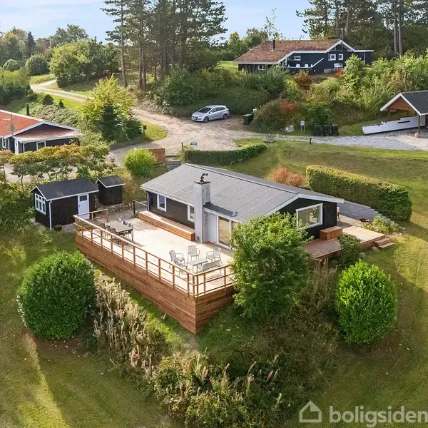 Et hus med træterrasse står på en frodig græsplæne omgivet af buske og træer. Nærliggende er der flere små huse og veje.