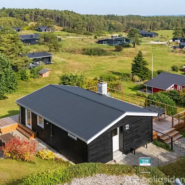 Sort sommerhus med nummer 12 på væggen, omgivet af grønne buske og græsplæne i et naturskønt område med spredte huse. En informationsplade er synlig ved indgangen.