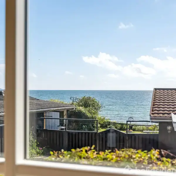 Havets blå bølger ses gennem et vindue, mens solen skinner over sommerhuse og en have i forgrunden.