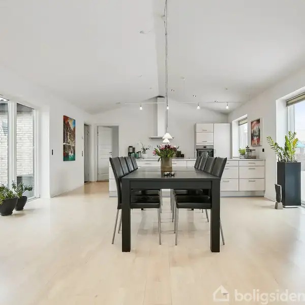 Spisebord med sorte stole står midt i et lyst køkken-alrum. Rummet har lyse trægulve, store vinduer, planter og moderne kunst på væggene. Køkkenet er i baggrunden.