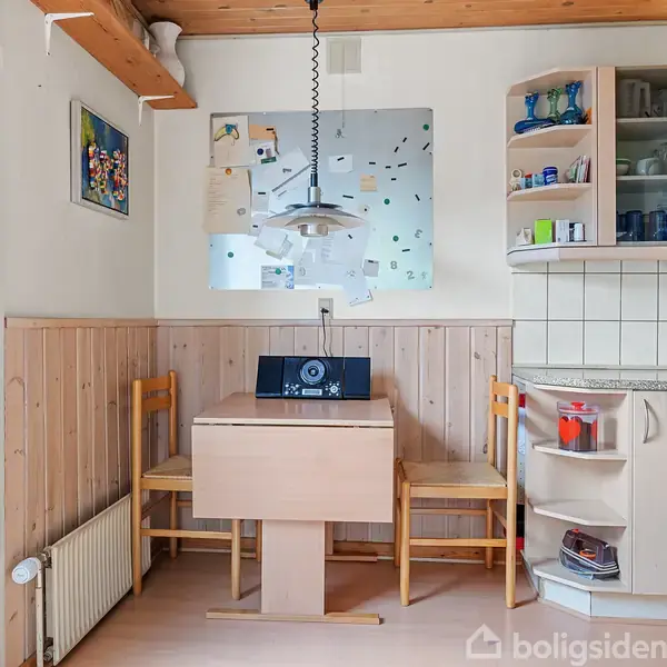 Et lille køkkenhjørne med et lyst træbord omgivet af to stole. En retro radio står på bordet. Over bordet hænger en lampe. Væggen bagved har hylder med glas og kopper.