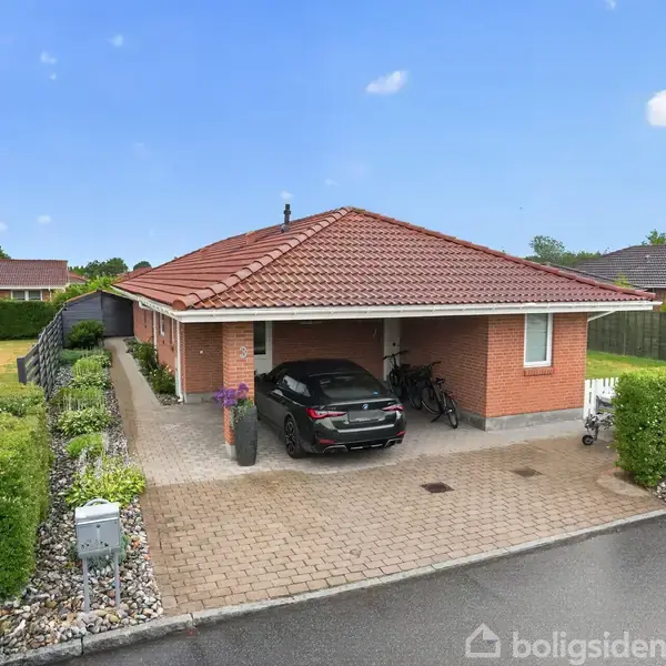 En murstensvilla med rødt tegltag har en sort bil parkeret i carporten. Villaen er omkranset af hække, og en cykel står ved siden af bilen. Op ad huset er en terrasse med blomsterkrukke.