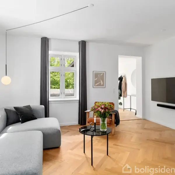 En grå sofa står i en stue, flankeret af et sofabord med blomster. Værelset har en stor vindue med gardiner, en vægmonteret fladskærm, og lyst trægulv.