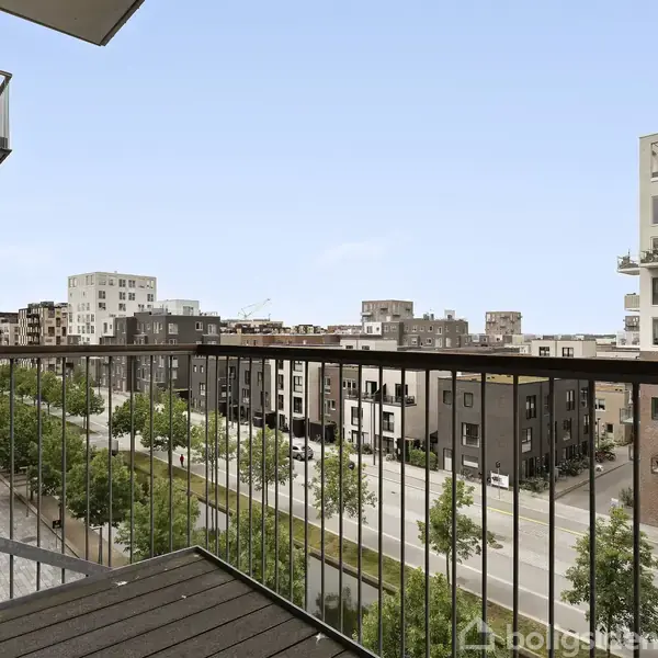 En balkon giver udsigt over en by, med moderne lejlighedsbygninger og en vej med træer. Himlen er klar og blå.