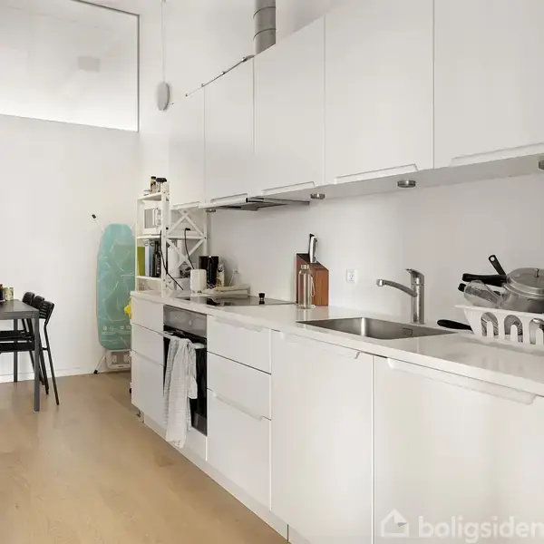 Hvidt køkken har lange skabe mod væg genstande på bordpladen, herunder skærebakker og service. Modsat er spisebord med sorte stole i et lyst rum.