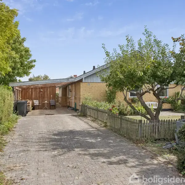 En bungalow med gult murværk ligger for enden af en flisebelagt indkørsel. Omringet af buske og træer, ses en træterrasse ved siden af huset.