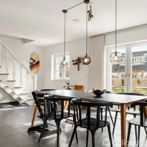 Et moderne spisebord med sorte stole står centralt i et lyst rum. Ved siden af er en hvid trappe, og store vinduer viser en udendørs terrasse. En plante står nær trappen.