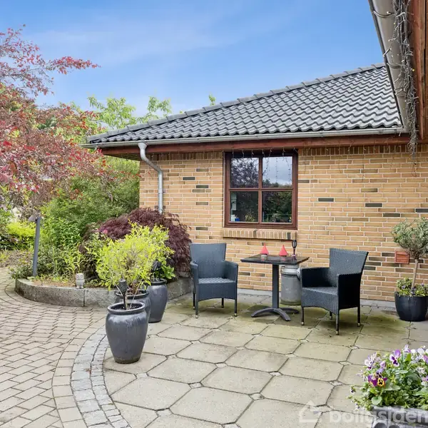 Terrasse indrettet med to sorte stole og et rundt bord. Omgivet af plantebeholdere med buske og blomster op ad en gul murstensbygning med gråt tag.