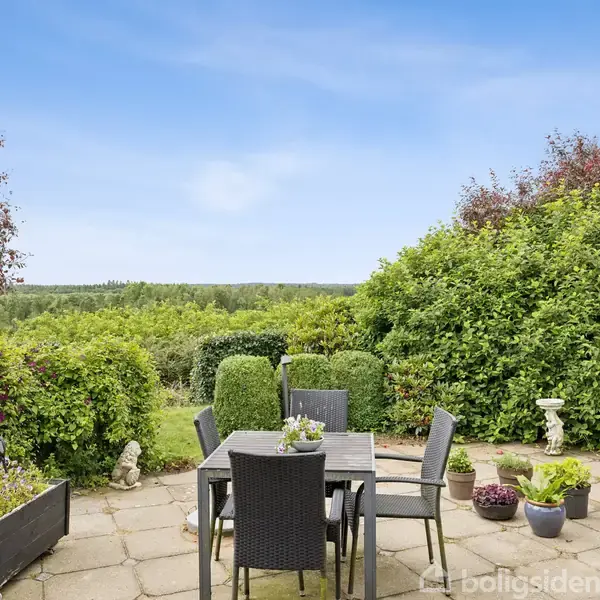 Sort havemøblement står på en flisebelagt terrasse omgivet af grønne buske og blomsterkrukker. I baggrunden ses en frodig, grøn udsigt under en klar blå himmel.