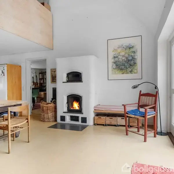 Pejs med ild brænder i en lys stue. Til venstre står et spisebord og stole, og der er en dør til en terrasse. Maleri hænger ved siden af pejsen.