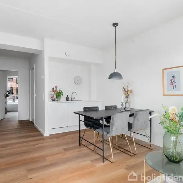 Spisebord med fire stole står på trægulv i et lyst køkken-alrum. Bordet er dekoreret med en vase med blomster, og tre billeder hænger på væggen.