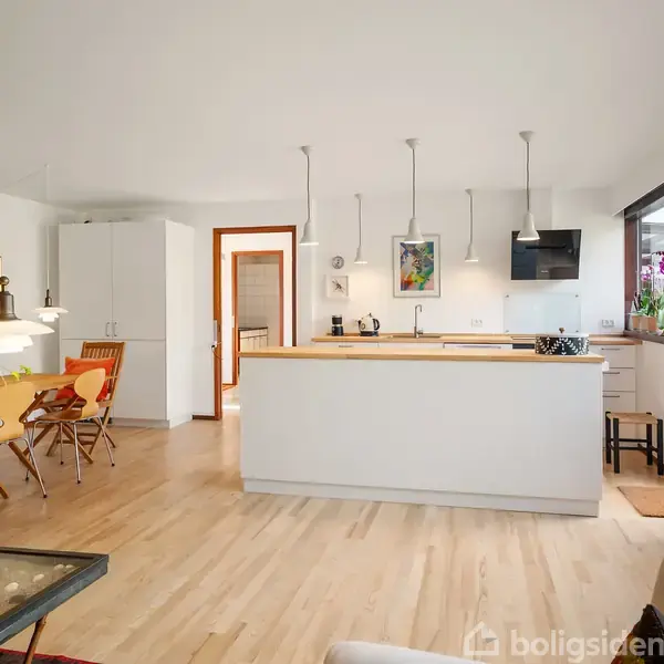 Køkken-alrum med hvidt køkkenø, tre hængelamper, spisebord med stole til venstre. Store vinduer til højre giver lys, mens planter og dekorationer pryder rummet.