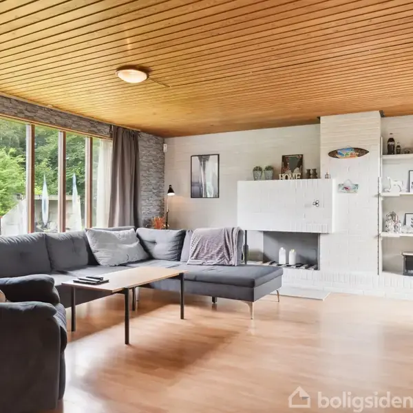 En grå hjørnesofa står ved et lavt sofabord i en lys stue med trægulve og træloft. Store vinduer og grå gardiner slipper dagslys ind. En hvid pejs og hylder fyldt med dekorationer ses ved siden af.