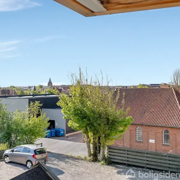 Biler holder parkeret på en grusplads ved siden af en bygning med rødt tag. Træer omkranser området, og en by ses i baggrunden under en klar blå himmel.