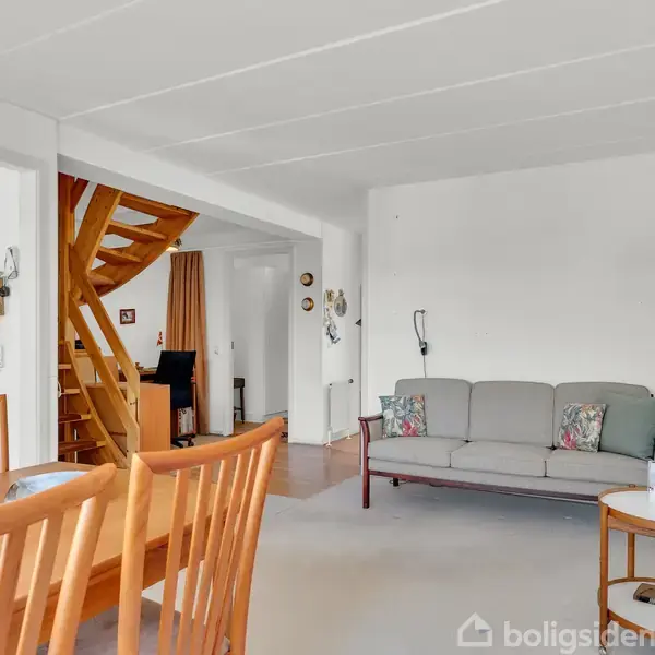 En sofa står i en stue med et lyst interiør. Et spisebord med stole er i forgrunden, og en trætrappe leder til overetagen. Baggrunden viser en åben rumfordeling.