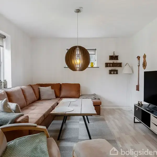 Brunt hjørnesofa-arrangement med puder står i en lys stue. Foran står et sofabord, og til højre ses et tv på et lavt skab. En moderne lampe hænger over bordet.