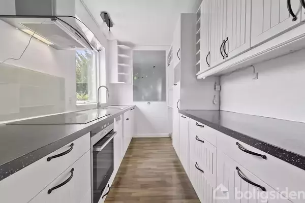 Køkken med hvide skabe og sorte bordplader, indeholder komfur og vask. Rummet har trægulv og et stort vindue, der giver lyse omgivelser.