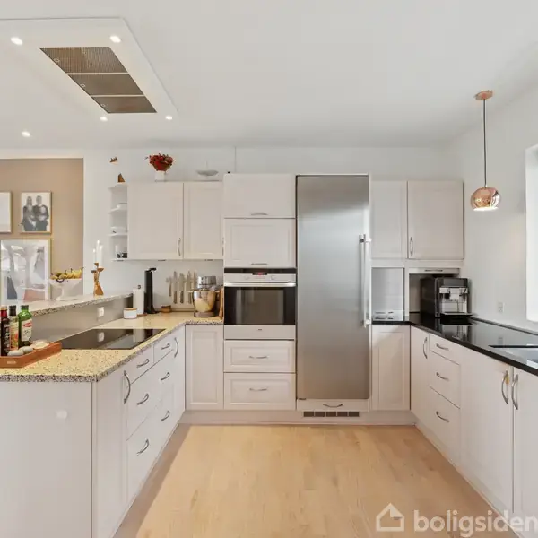 Et moderne køkken med hvide skabe og en stål køleskab i centrum. Køkkenet har en sort granitbordplade, lys trægulv og vindue mod højre. Flere billeder hænger på væggen.