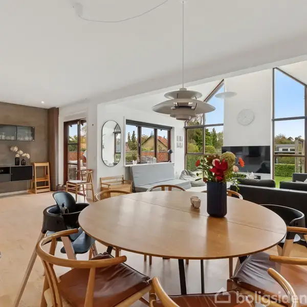 Rundt spisebord med stole står i et moderne, lyst rum med hvide vægge og store vinduer. I baggrunden ses et åbent køkken og en stue med grå sofaer og et tv.