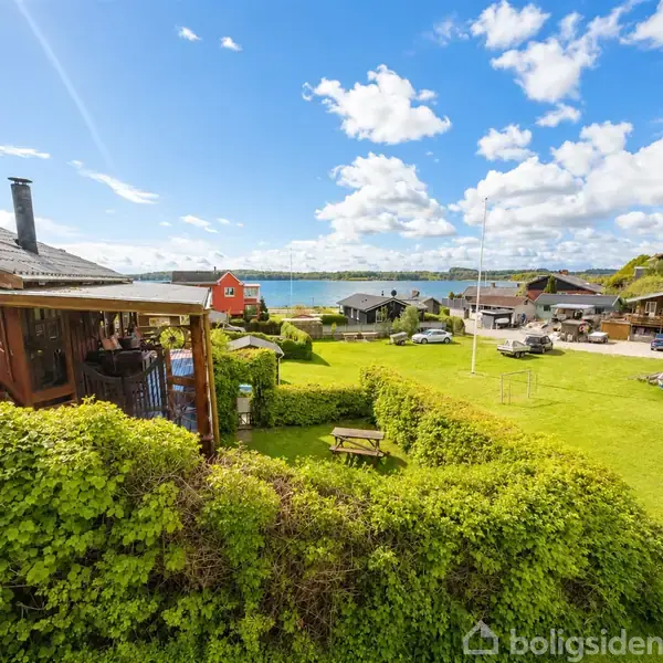 Et hus med veranda står stille blandt grønt græs og buske. I baggrunden ses flere huse og en sø i sollys. Skyer fylder den blå himmel.