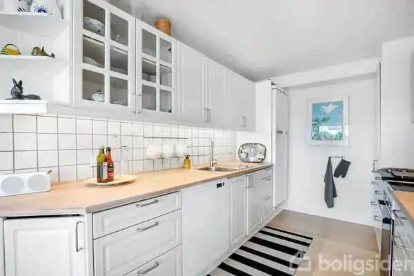 Et hvidt køkken med skabe, køkkenbord fyldt med olieflasker og en skål, stribet tæppe på gulvet, fliser på væggen og håndklæde; moderne og ryddeligt.