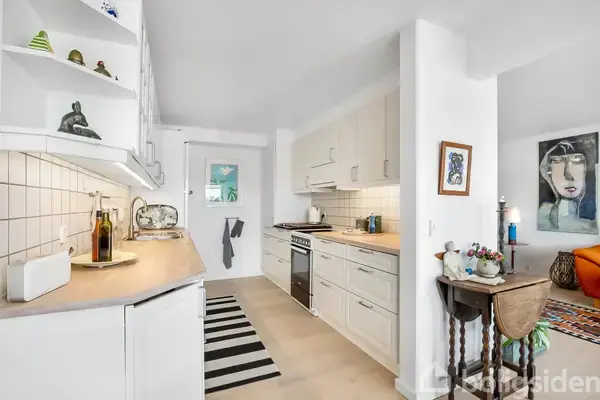 Et lyst køkken med hvide skabe og træbordplader. En stribet løber ligger på gulvet. Ved siden af køkkenet ses en stue med farverige møbler og kunst på væggene.