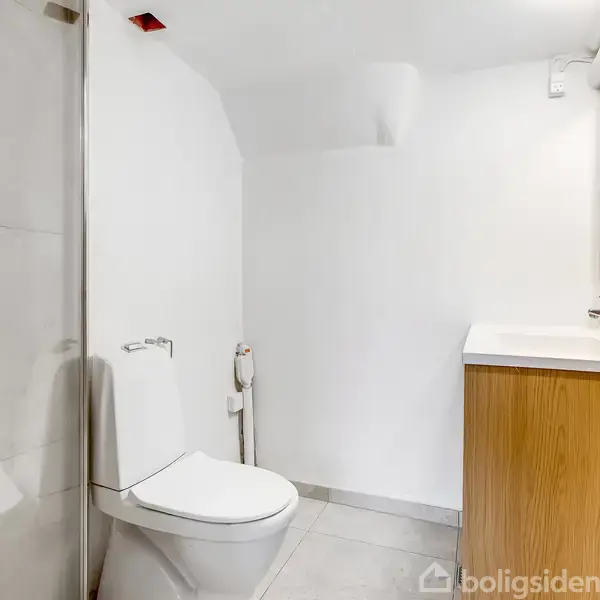 Toilet placeret ved siden af en bruseniche, omgivet af hvide fliser og vægge. En vask med et spejl hænger ved siden af i et lyst og enkelt badeværelse.