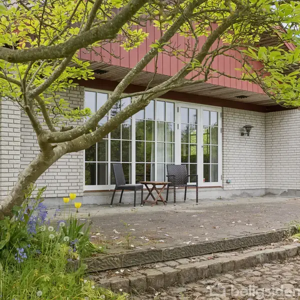Et hus med hvid murstensfacade står bag en terrasse, hvor to stole og et bord er placeret. Omgivet af grønne planter og træer i en frodig have.