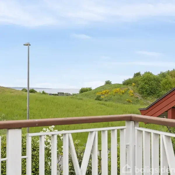 Rækværk omgiver en balkon, der er dækket af træbrædder, med udsigt over en bakke med grønt græs og buske. En lygtepæl står i baggrunden mod en blå himmel.