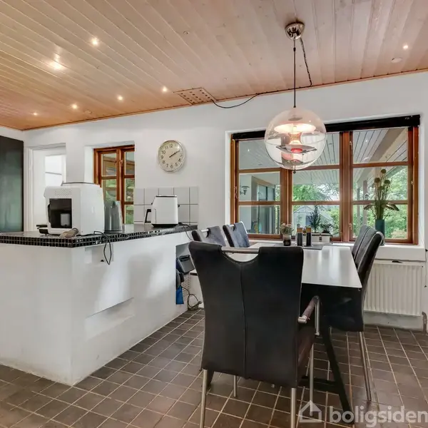 Et moderne køkken-alrum med spisebord og sorte læderstole. Et køkkenbord med apparater står centralt. Store vinduer giver udsigt til en terrasse. Loftslamper belyser rummet. Vægur viser tid.
